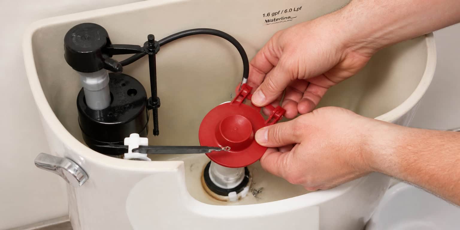 A close-up photo of hands fixing a running toilet, replacing the red toilet flapper inside the ceramic tank, bright bathroom lighting.